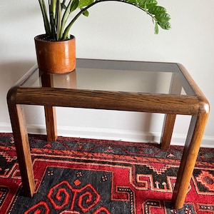 May include: A mid-century modern side table with a rectangular glass top and a wooden frame. The table is approximately 24 inches long, 18 inches wide, and 20 inches tall. A potted plant sits on the table.