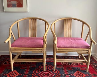 May include: Two bamboo armchairs with curved backs and pink cushions. The chairs have a natural wood finish and are placed on a patterned rug with red and dark blue tones. The chairs are positioned in front of a white wall.
