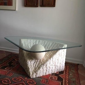 May include: A modern coffee table with a clear glass top and a textured, off-white stone base. The base features a spherical stone element. The table sits on a patterned rug.