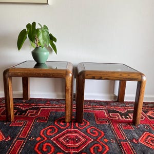 May include: Two vintage wooden side tables with glass tops. The tables have a warm, medium-brown finish and rounded corners. One table has a green vase with a plant on top. The tables are on a patterned rug.