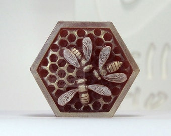 Könnte beinhalten: Eine sechseckige Seife mit einem Wabenmuster und drei Bienen in Relief. Die Bienen sind silber und gold, und die Wabe ist braun und gold.