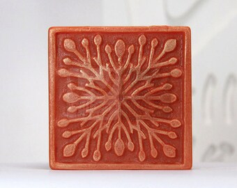 Könnte beinhalten: Eine quadratische, terrakottafarbene Seife mit einem erhabenen, filigranen Design einer stilisierten Blume in Gold.