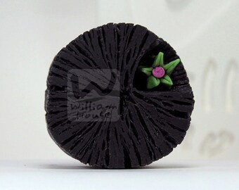 Könnte beinhalten: Eine schwarze, runde Holzscheibe mit einer grünen und pinken Blume darauf. Die Scheibe hat eine Holzmaserung.