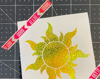 May include: A gold glitter sun with a yellow centre on a white background. The sun has a textured, sparkly finish. The background is a black cutting mat with white grid lines.