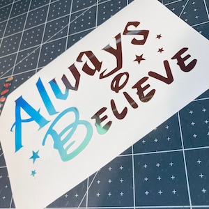 Pode incluir: Um adesivo de vinil branco com as palavras "Always Believe" em uma fonte degradê azul e marrom. As palavras estão em uma fonte estilizada com estrelas ao redor.