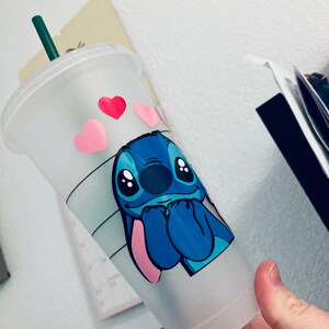 May include: A white reusable cup with a blue Stitch character from Lilo & Stitch. Stitch is holding his hands together and has a pink nose. There are pink and red hearts around Stitch.