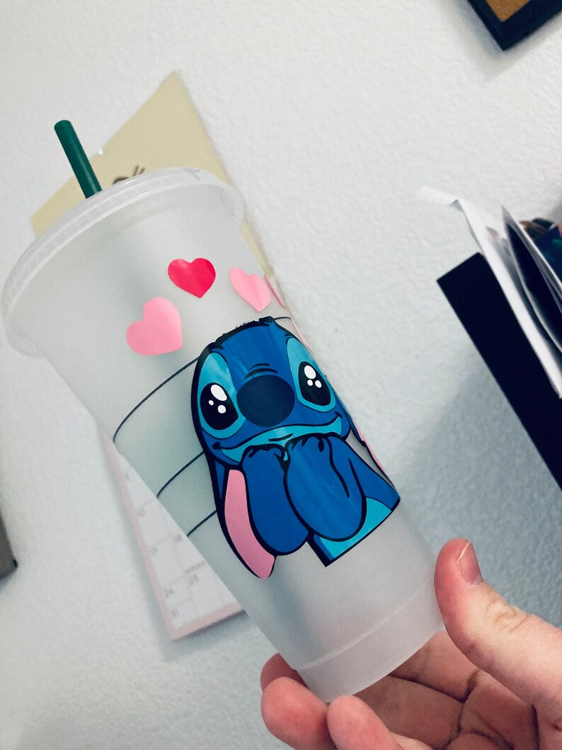 May include: A white reusable cup with a blue Stitch character from Lilo & Stitch. Stitch is holding his hands together and has a pink nose. There are pink and red hearts around Stitch.