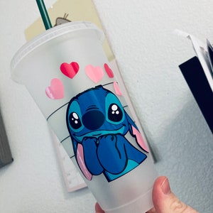 May include: A clear plastic reusable cup with a blue Stitch character from Lilo & Stitch. The cup has a green straw and pink hearts around the top.