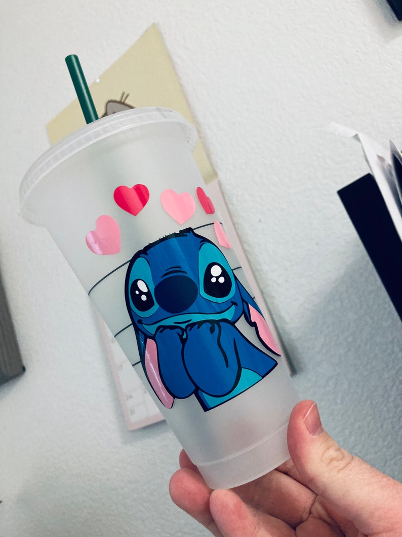 May include: A clear plastic reusable cup with a blue Stitch character from Lilo & Stitch. The cup has a green straw and pink hearts around the top.