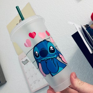 May include: A frosted clear plastic cup with a blue cartoon character with pink ears and big eyes. The character is holding its hands together in front of its face. The cup has pink hearts and a black and white striped pattern around the bottom.