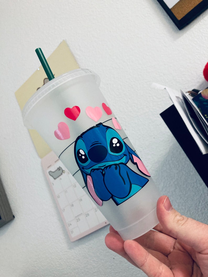 May include: A frosted clear plastic cup with a blue cartoon character with pink ears and big eyes. The character is holding its hands together in front of its face. The cup has pink hearts and a black and white striped pattern around the bottom.