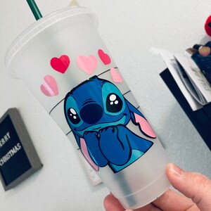 May include: A clear plastic reusable cup with a blue Stitch character from Lilo & Stitch. Stitch is holding his hands up to his face and has pink hearts around his head. The cup has a green straw.