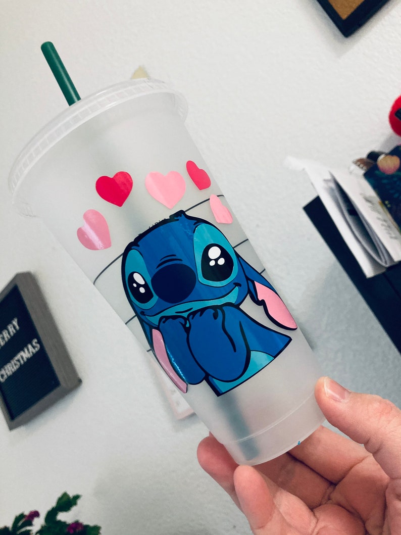 May include: A clear plastic reusable cup with a blue Stitch character from Lilo & Stitch. Stitch is holding his hands up to his face and has pink hearts around his head. The cup has a green straw.
