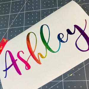 Könnte beinhalten: Ein regenbogenfarbener, schimmernder Vinyl-Aufkleber mit dem Namen "Ashley" in kursivschrift.
