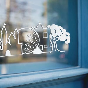 May include: White vinyl decal featuring a stylized outline of a castle, a geometric sphere, and a tree. The decal is applied to a window, with a blue window frame in the background. The design is a silhouette.