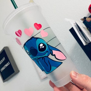 May include: A clear plastic reusable cup with a blue cartoon character with pink ears and big eyes. The character is winking and has pink hearts around it. The cup has a green straw.