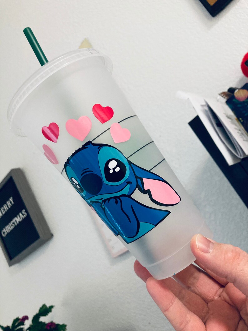May include: A clear plastic reusable cup with a blue cartoon character with pink ears and big eyes. The character is winking and has pink hearts around it. The cup has a green straw.