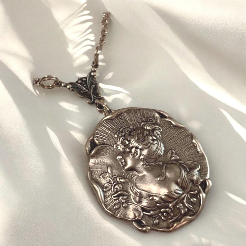 May include: Silver-toned pendant necklace featuring a detailed relief of a woman's profile. The oval pendant has an ornate frame and is suspended from a delicate chain. The woman's profile is surrounded by a sunburst design.