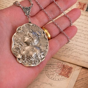 May include: A silver-toned pendant necklace featuring a detailed cameo design of a woman in profile, holding a flower. The pendant is attached to a delicate chain. The necklace is held in a hand, with vintage letters in the background.