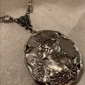 May include: A silver-toned pendant necklace featuring a detailed cameo of a woman's profile. The oval pendant is intricately designed with floral and sunburst elements, suspended from a delicate chain.