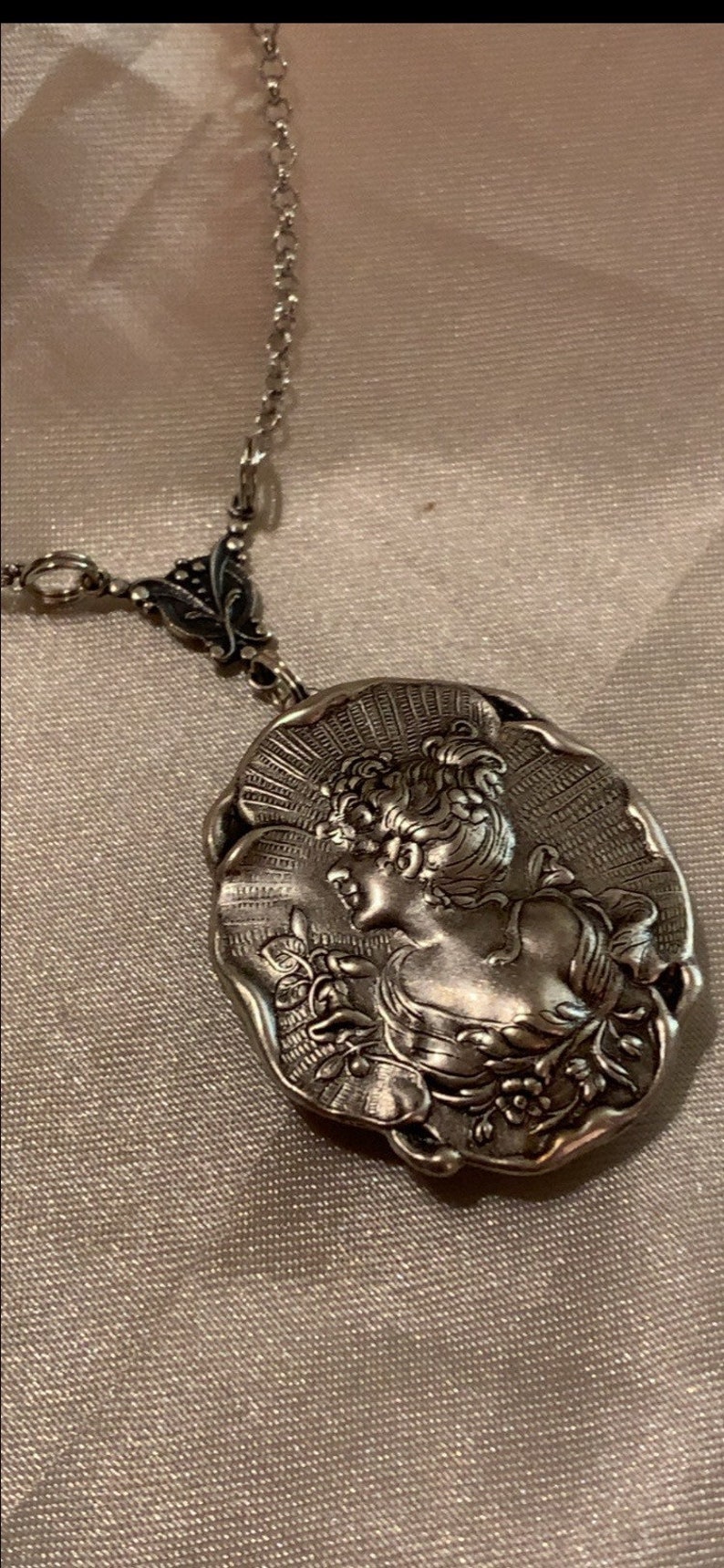 May include: A silver-toned pendant necklace featuring a detailed cameo of a woman's profile. The oval pendant is intricately designed with floral and sunburst elements, suspended from a delicate chain.