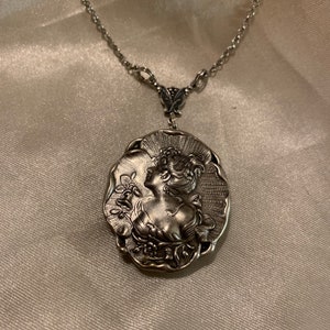May include: Silver-toned pendant necklace featuring a detailed relief of a woman's profile with floral accents. The oval pendant is suspended from a delicate chain. The necklace is displayed on a satin-like surface.