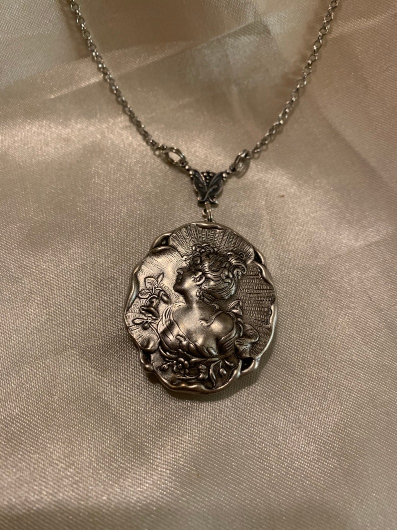 May include: Silver-toned pendant necklace featuring a detailed relief of a woman's profile with floral accents. The oval pendant is suspended from a delicate chain. The necklace is displayed on a satin-like surface.