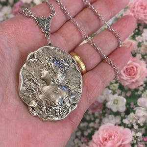 May include: A silver-toned pendant necklace featuring a detailed relief of a woman's profile within a floral frame. The pendant is suspended from a delicate chain. The background is a soft focus of pink and white flowers.