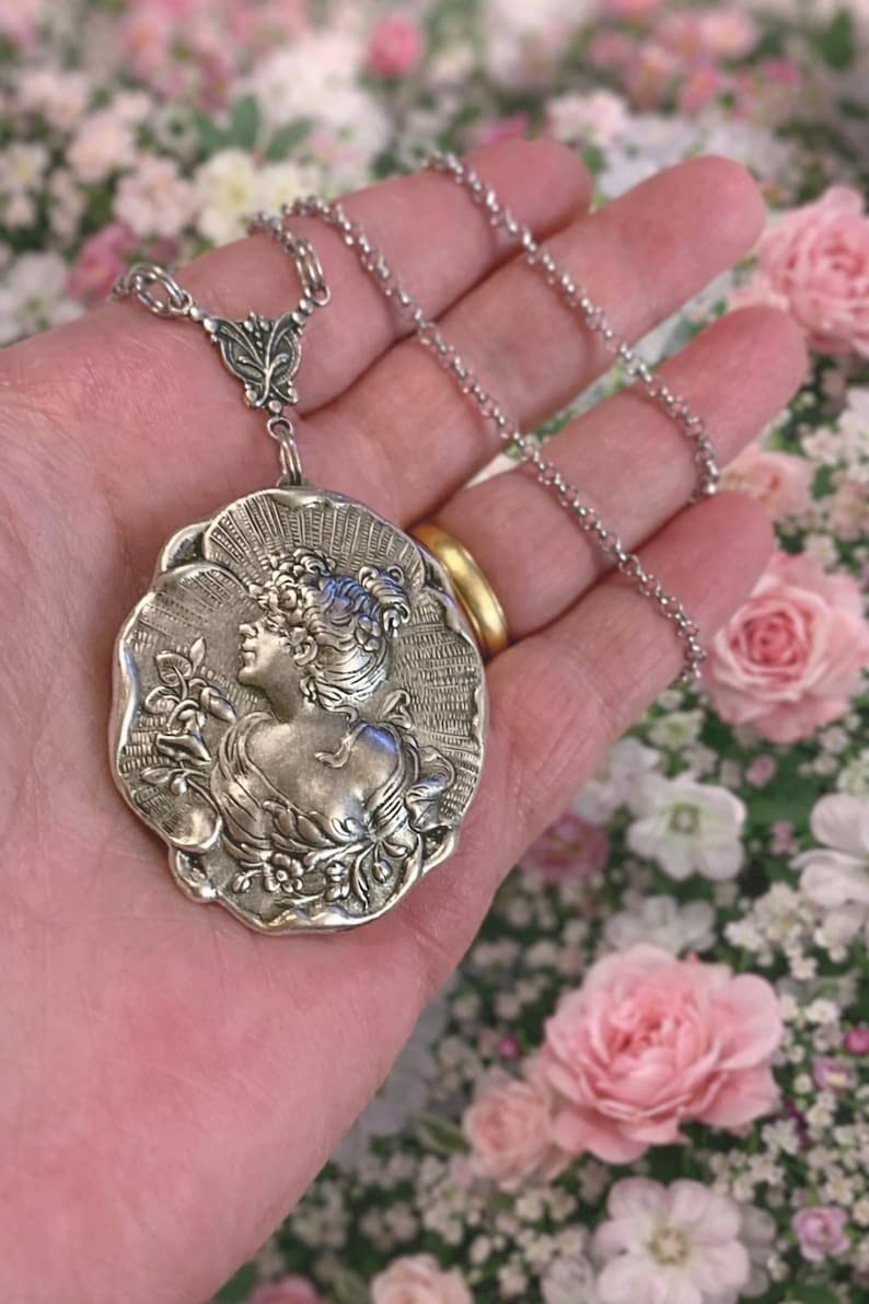 May include: A silver-toned pendant necklace featuring a detailed relief of a woman's profile within a floral frame. The pendant is suspended from a delicate chain. The background is a soft focus of pink and white flowers.