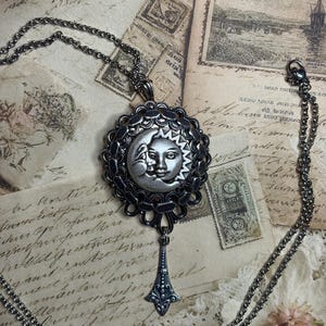 May include: A silver-toned celestial pendant necklace. The pendant features a sun and moon design with intricate detailing. The necklace has a delicate chain and a decorative drop. The background includes vintage ephemera.