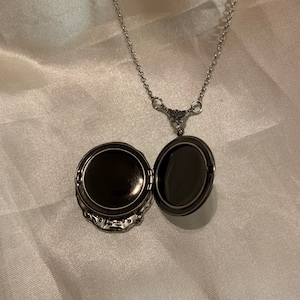May include: Open silver locket necklace with a decorative floral design. The locket is open, revealing a dark interior. The chain is delicate and hangs on a white satin background.