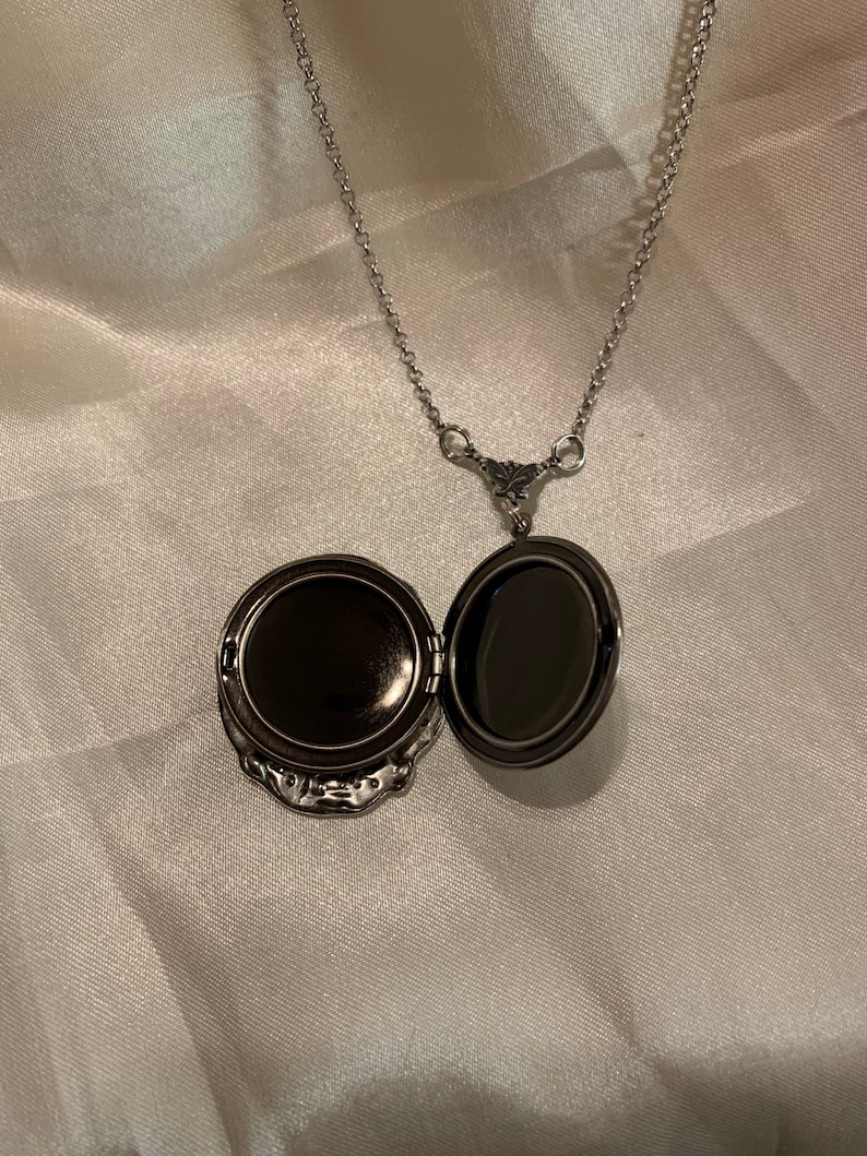 May include: Open silver locket necklace with a decorative floral design. The locket is open, revealing a dark interior. The chain is delicate and hangs on a white satin background.