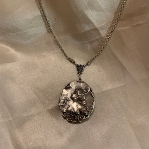May include: Silver-toned pendant necklace featuring an oval pendant with a detailed relief of a woman in profile. The pendant is suspended from a delicate chain. The necklace is a vintage-inspired piece.