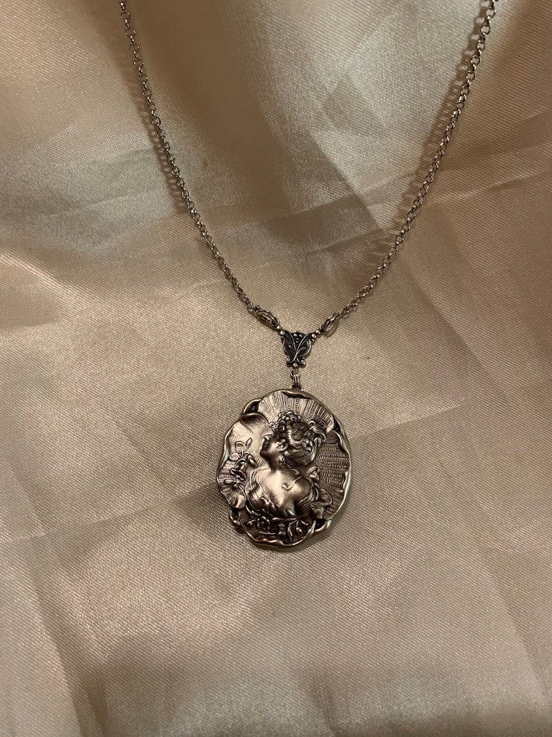 May include: Silver-toned pendant necklace featuring an oval pendant with a detailed relief of a woman in profile. The pendant is suspended from a delicate chain. The necklace is a vintage-inspired piece.