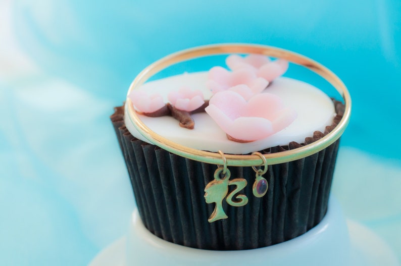 May include: A chocolate cupcake topped with white icing and pink flower decorations. A gold bracelet with a charm is placed on top of the cupcake.