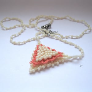 Può includere: Una collana con un ciondolo triangolare realizzato con perline color crema e un bordo in filo color corallo. La catenina è composta da un delicato filo di piccole perle color crema e include una chiusura a moschettone in metallo argentato.