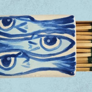 May include: A white ceramic matchbox with a blue and white design of three fish with large eyes. The matchbox is filled with wooden matches with green tips.
