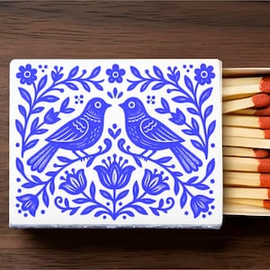 May include: A rectangular matchbox featuring a white cover with a blue illustration of two birds facing each other, surrounded by floral and leaf patterns. The sliding tray is partially open, revealing a row of wooden matches with orange tips.