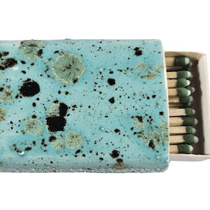 May include: A teal blue matchbox with a speckled pattern. The box is open and shows a stack of matches with green tips.