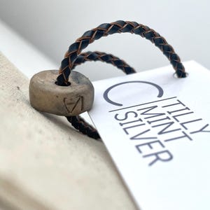 May include: A braided black leather cord loop threaded through a cylindrical tan ceramic bead with an etched letter. A white rectangular paper tag is attached to the cord, featuring the printed text TILLY MINT SILVER in black lettering.
