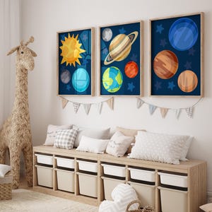May include: Three framed art prints featuring colorful illustrations of planets and the sun against a dark blue starry background. The artwork is displayed above a wooden storage bench with white bins, pillows, and a decorative giraffe.