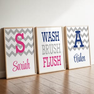 May include: Three framed rectangular wall art prints featuring a gray and white chevron pattern. The first print displays a pink letter S and the name Sariah. The second print features the words WASH, BRUSH, and FLUSH. The third print shows a blue letter A and the name Aiden.