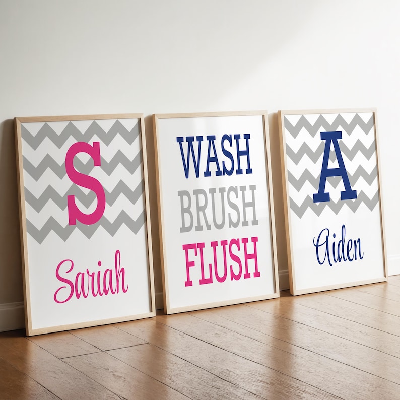 May include: Three framed rectangular wall art prints featuring a gray and white chevron pattern. The first print displays a pink letter S and the name Sariah. The second print features the words WASH, BRUSH, and FLUSH. The third print shows a blue letter A and the name Aiden.