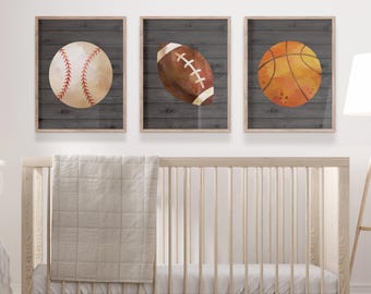 May include: Three framed prints with a wood-grain background. Each print features a different sports ball: a baseball, a football, and a basketball. The balls are painted in a watercolor style.
