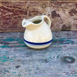 Könnte beinhalten: Ein weißer Keramik-Krug mit einem blauen Streifen um die Mitte. Der Krug hat einen Henkel und einen Ausguss. Er steht auf einer Holzoberfläche.