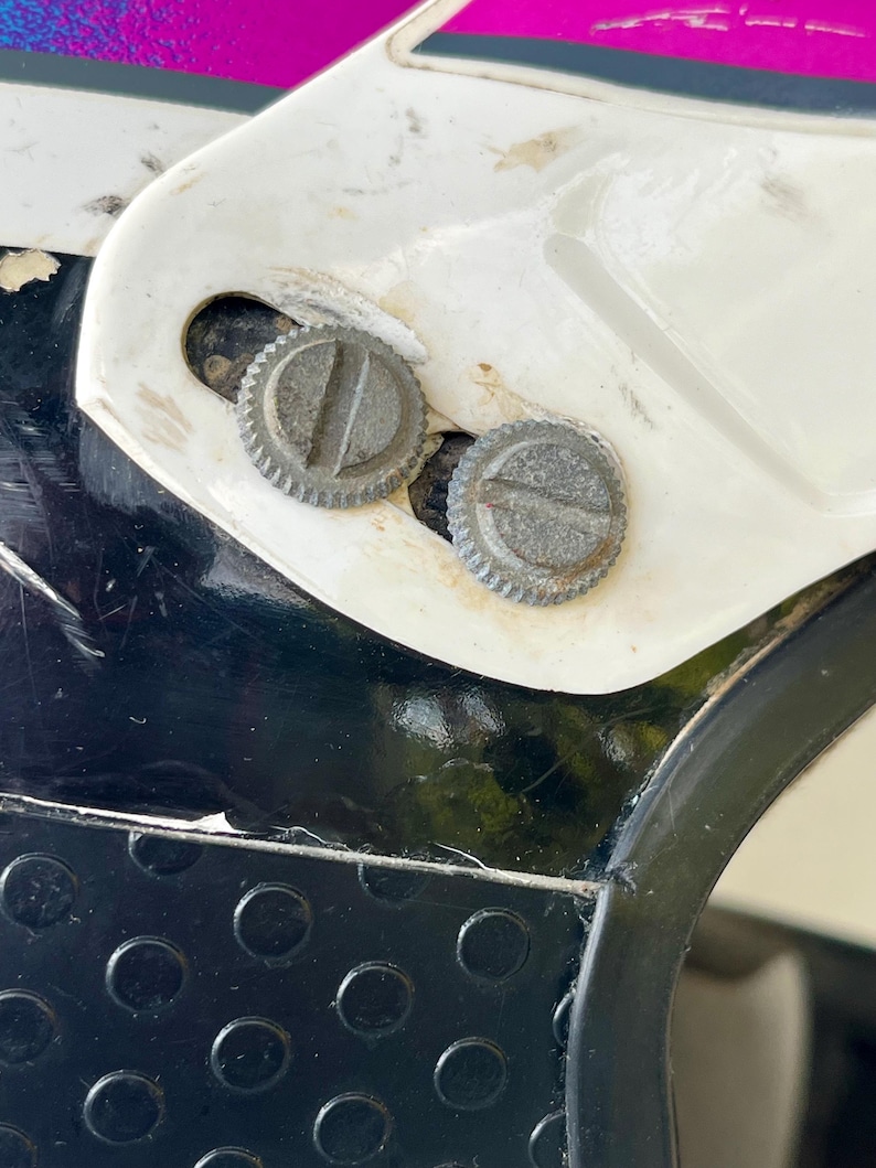 May include: Close-up of a section of a white and black motorcycle helmet. Two silver, circular screws are visible, set into the white plastic. The helmet has a black, textured section with circular indentations.