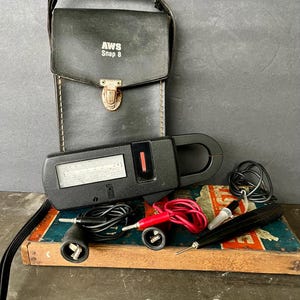 May include: A black AWS Snap 8 clamp meter with a leather carrying case. The meter has a digital display and comes with test leads and a probe. The case is black with a buckle closure.