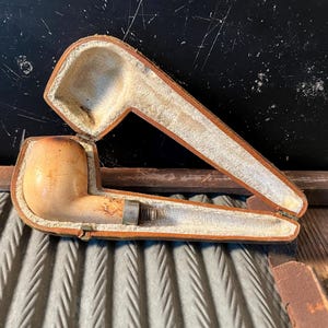 May include: A vintage tobacco pipe with a light-colored bowl and a metal stem, resting inside its open, rectangular case. The case is lined with a soft, off-white material and has a brown leather exterior.