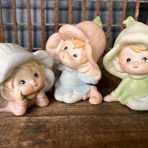 May include: Three ceramic figurines of flower fairies in pastel colors. Each fairy has a flower hat and is in a different pose. The figurines are on a wooden surface with a metal grate background.