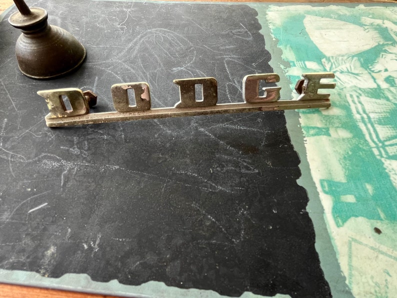 May include: A silver metal car emblem with the word "DODGE" in raised letters.